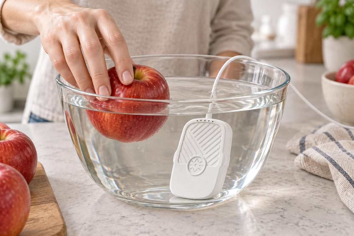A hand placing an apple in a glass bowl of water beside the white FreshDip cleaner.